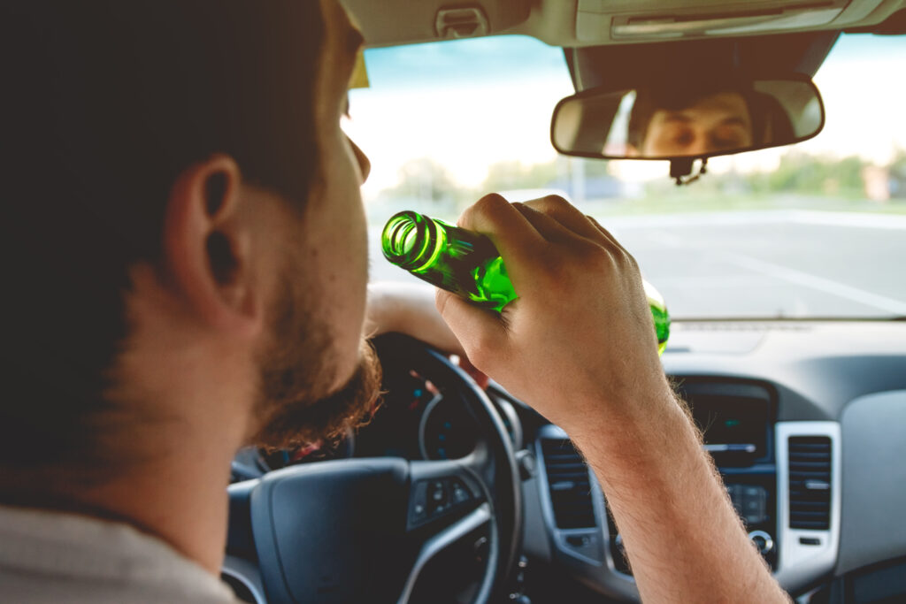 A young man with a beard is sitting in the driver's seat of a car, holding a green glass bottle close to his mouth. His right hand grips the steering wheel while his left hand holds the bottle. His reflection is visible in the rearview mirror. The car's interior, including the dashboard and steering wheel, is clearly visible, and the background outside the windshield shows an open road with some greenery.