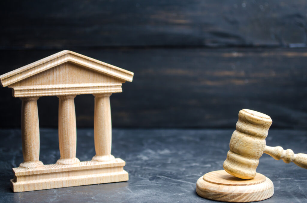 A wooden model of a classical building with columns, resembling a courthouse, is positioned next to a wooden gavel resting on its base. Both objects are placed on a dark, textured surface, symbolizing justice and legal authority.