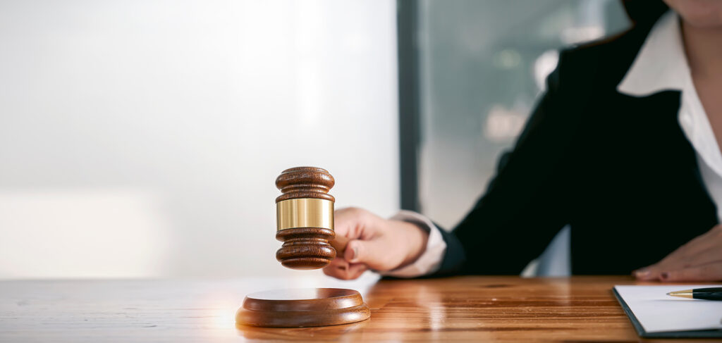 A judge or legal professional wearing a black suit and white shirt is about to strike a wooden gavel on a round sound block. The gavel is slightly above the block, appearing as if it is in motion. The setting is a courtroom or office with a wooden table, a notepad, and a pen visible in the foreground. The background is blurred, emphasizing the gavel and the person’s hand.