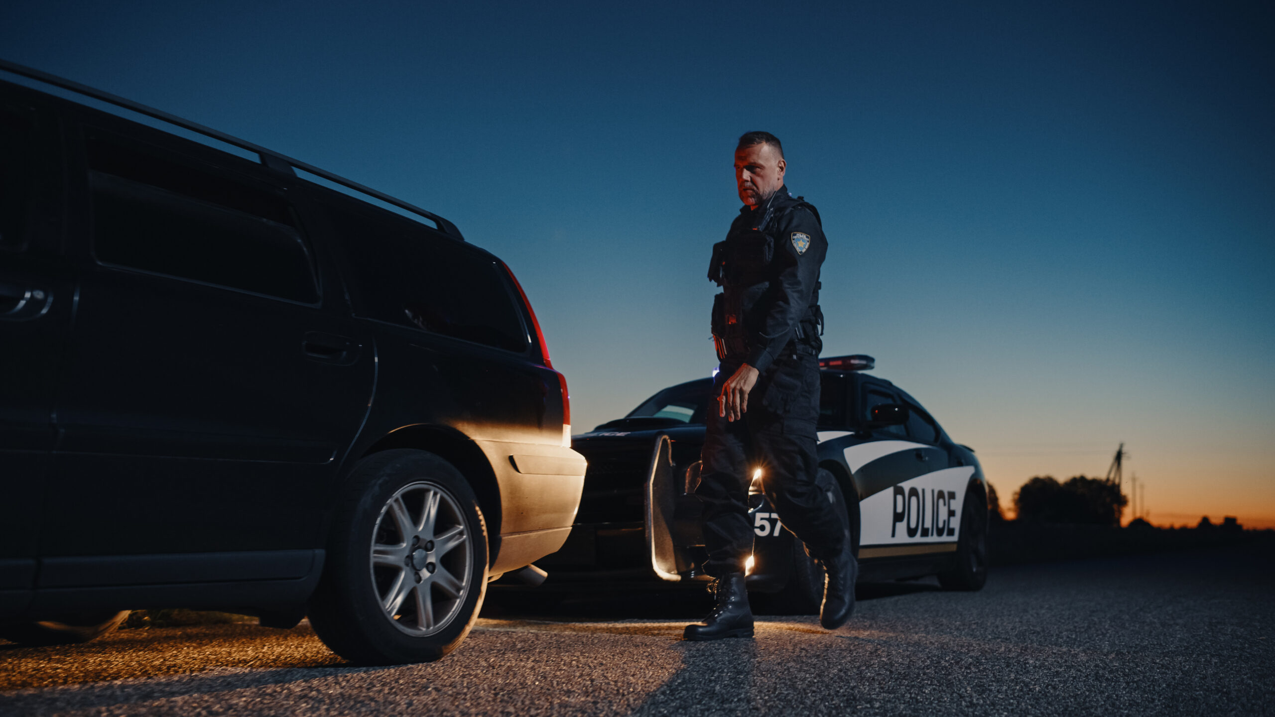 A police officer in a dark uniform walks towards a black SUV that has been pulled over on the side of the road during twilight. His expression is serious and focused, and he wears tactical gear with a badge on his sleeve. Behind him, a police car with flashing red and blue lights is parked, illuminating the scene. The background features a clear evening sky transitioning from deep blue to warm orange near the horizon, with silhouettes of trees and power lines visible in the distance. The atmosphere conveys authority and tension.