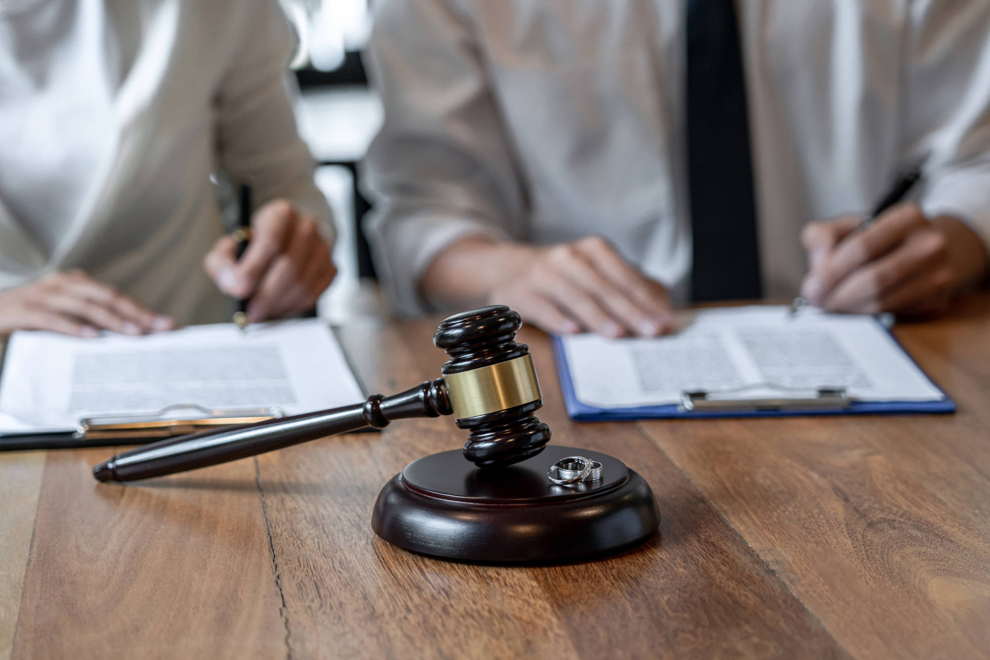 Close-up of a wooden table in a courtroom or legal office setting. Two people, likely lawyers or legal professionals, are seated at the table. They are both dressed in light-colored, professional attire, one is in a white collared shirt and tie, the other one in a similar light-colored shirt. Several documents are visible on the table, held by a metal binder clip. The papers appear to be legal documents, possibly contracts or agreements, that the two individuals are reviewing and signing. A wooden gavel rests on the table, its base sitting on a dark wooden stand. A pair of wedding rings are near the gavel, laying on the table.