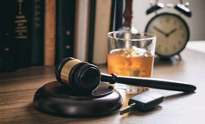 Close-up of a wooden gavel resting on a wooden stand, a glass of whiskey with ice, a car key, and an alarm clock in the background. The gavel, dark brown or black wood with a gold band around the head, is positioned centrally in the image. The stand it sits on is also dark wood, circular, and slightly larger than the gavel itself. A clear glass filled with amber-colored whiskey and ice cubes is situated beside and a bit behind the gavel, slightly tilted. A black alarm clock with silver numerals is in the upper-right of the image, out of focus. A car key is angled on the table, adjacent to or just below the glass of whiskey, also out of focus. Blurred books and legal-looking documents are in the background. The overall impression suggests a legal or courtroom setting, potentially involving discussion related to drinking and driving.