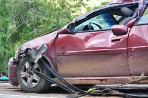 Close-up of a severely damaged red car. The car's front passenger side and a portion of the driver's side are heavily dented and crushed. Significant damage is visible to the body panels, the doors, and the front end. 